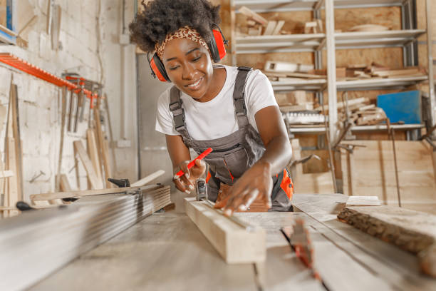 Woman Carpenter of African-American Ethnicity in Working Coverall is Preparing a Wooden Plank for Cutting. A Carpenter Measures Wooden Board with Measuring Tape While Working in a Workshop.